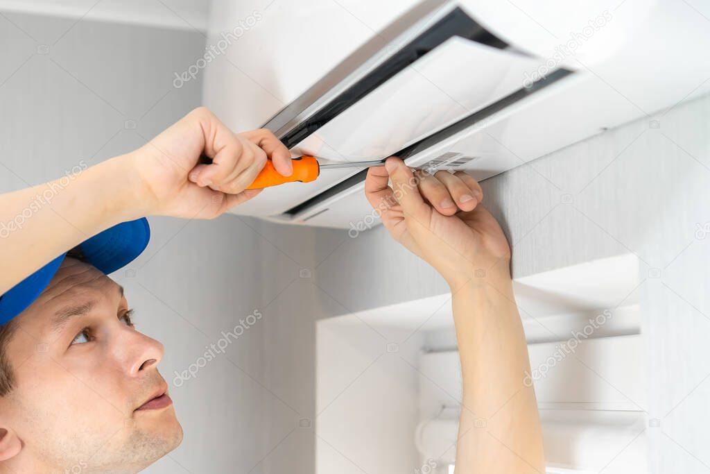 Repairman in overalls with a screwdriver in his hands repairs an air conditioner on the wall