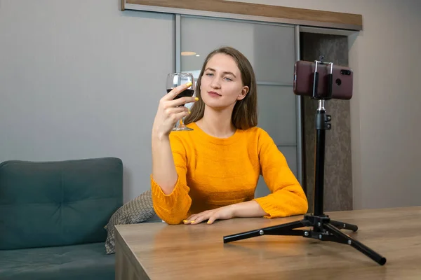 Young dark-haired female blogger in an orange sweatshirt with a glass of red wine sits in front of a tripod with a mobile phone in a stylish interior. Female wine blogger