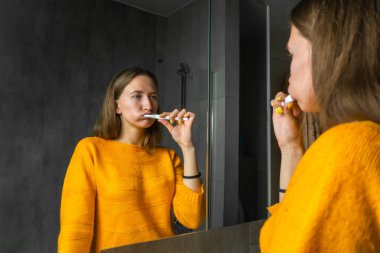 Turuncu kazaklı genç, koyu renk saçlı bir kadın sabah dişlerini gri tonlarda modern bir banyoda fırçalıyor.