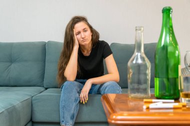 Genç siyah saçlı kadın kanepede oturuyor ve boş şişelerin ve sigaraların yanında üzgün üzgün bir bakışla kameraya bakıyor. Alkolizmin sosyal sorunu