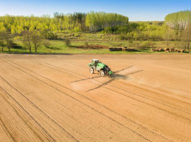 İlkbaharda, ormanın yakınındaki büyük bir arazide gübre ve kimyasalları püskürten bir traktörün drone görüntüsü.