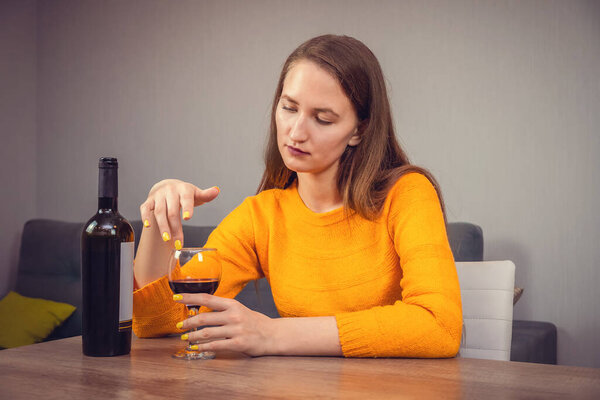 Close up thoughtful young dark-haired woman in an orange sweatshirt sits at the kitchen table, running her finger over a glass of red wine, bottle of wine in a dark glass next to it