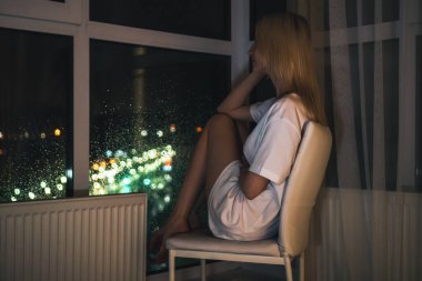 Beyaz gömlekli genç sarışın kadın çenesi yukarıda bir sandalyede oturur ve bir yerden bir tavana gece şehrini seyreder.