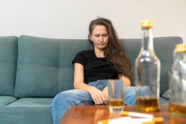 Genç siyah saçlı kadın gözleri kanepeye kapalı oturuyor, boş şişelerin ve sigaraların yanında. Alkolizmin sosyal sorunu