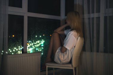 Beyaz gömlekli genç sarışın kadın çenesi yukarıda bir sandalyede oturur ve bir yerden bir tavana gece şehrini seyreder.