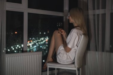 Beyaz tişörtlü, bir bardak kırmızı şaraplı genç bir sarışın bir sandalyeye oturur ve gece şehrine pencereden bakar. Seçici odak