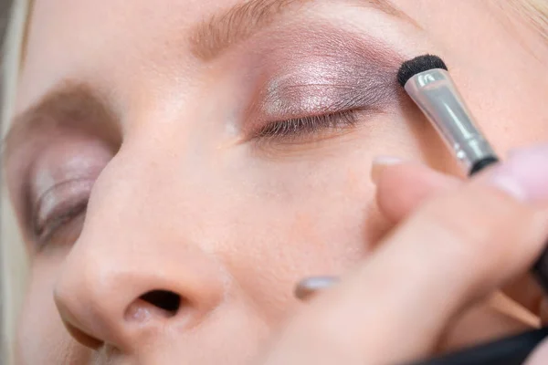 Make-up artist makes eye makeup for a young woman in brown and pink close-up