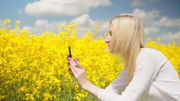 Hermosa mujer toma selfie en su teléfono inteligente en el campo de la floración de la colza. — Vídeos de Stock