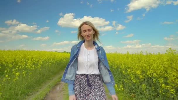 Μια όμορφη γυναίκα περπατάει μπροστά στην κάμερα μέσα από ένα χωράφι με κραμβόσπορους μια ηλιόλουστη μέρα. — Αρχείο Βίντεο