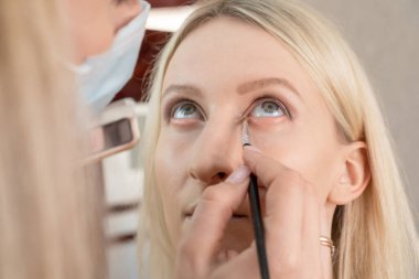 Makyaj sanatçısı, kahverengi ve pembe tonlu genç bir kadının gözlerini yakın plan çekiyor. Göz kapağının altındaki gölgeleri gölgeliyor.