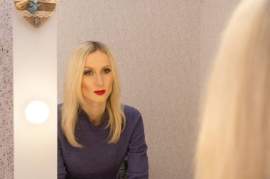 Dudağında kırmızı ruj lekesi olan sarışın, genç ve güzel bir kadın aynada makyajcının yaptığı işi inceliyor.