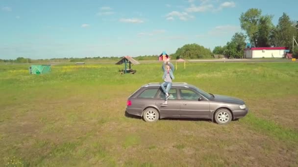Shooting fra drone af bil, på taget af som sidder en ung kvinde i en denim dragt. – Stock-video