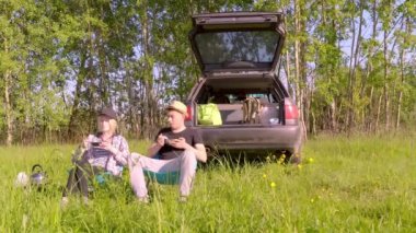 Genç adam ve kadın doğadaki sandalyelerde oturur ve arabanın yanındaki çayırlarda yemek yerler.