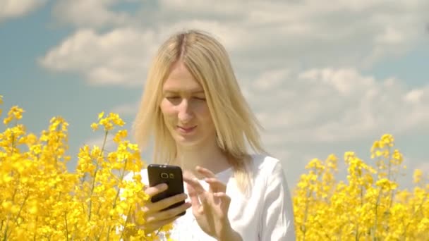 Joven hermosa rubia toma fotos de una colza en flor en un campo — Vídeo de stock