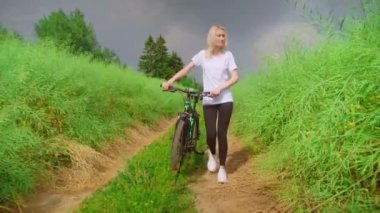 Bir kız bisikletle bir arazi yolunda yürüyor. taytlı ve beyaz gömlekli.