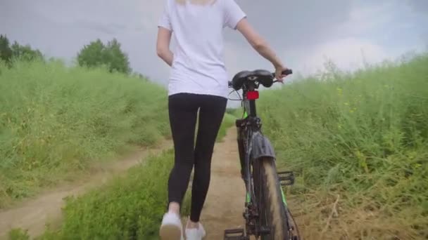 Uma menina está andando ao longo de uma estrada de campo com bicicleta. em leggings e camisa branca — Vídeo de Stock