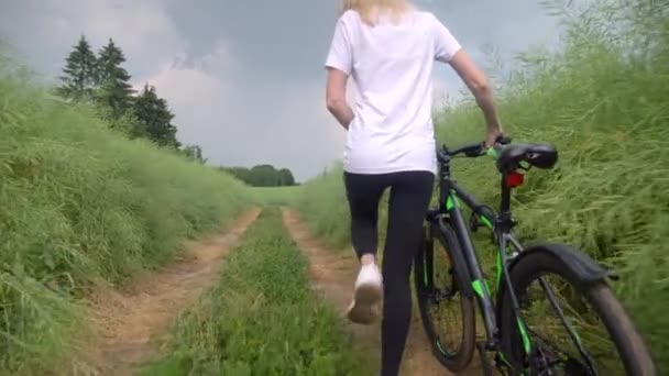 Uma menina está andando ao longo de uma estrada de campo com bicicleta. em leggings e camisa branca — Vídeo de Stock