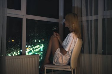 Beyaz tişörtlü, bir bardak kırmızı şaraplı genç bir sarışın bir sandalyeye oturur ve gece şehrine pencereden bakar. Seçici odak