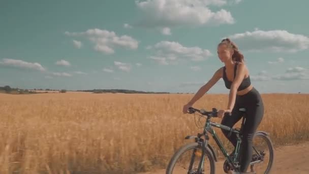 Uma jovem mulher monta uma bicicleta em uma estrada rural em um campo em um dia ensolarado. — Vídeo de Stock