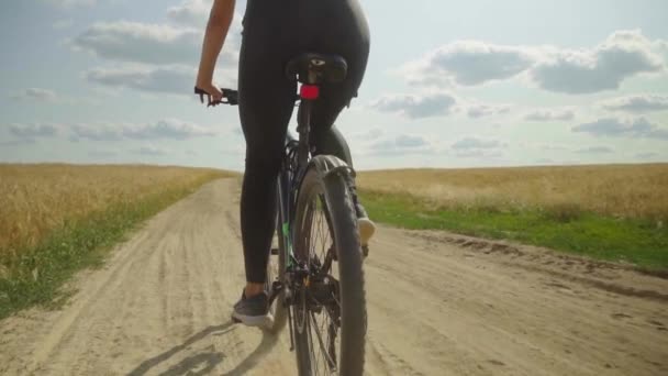 Uma jovem mulher monta uma bicicleta em uma estrada rural em um campo em um dia ensolarado. — Vídeo de Stock