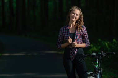 Güneşli bir yaz gününde, tişörtlü, taytlı ve bisikletli gülümseyen genç bir kadının portresi.