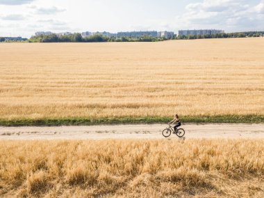 Güneşli bir yaz gününde trikalle tarlanın ortasında bisiklet süren genç bir kadının uzaktan kumandalı görüntüsü, arka planda şehir evleri görülüyor. Yan görünüm.