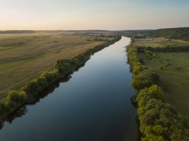 Nehrin insansız hava aracı manzarası, ağaçlar, çalılar, nehir boyunca kumlu bir yol bir yaz akşamı gün batımında