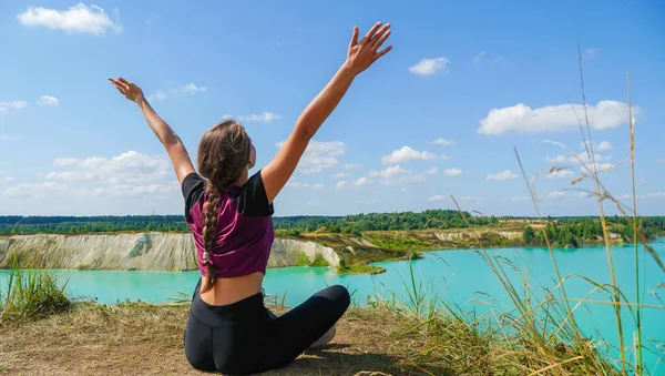 Young woman in leggings and a top with her hands raised up sits in a lotus position on the edge of a chalk quarry on a sunny summer day, Belarus. Concept of freedom, sport