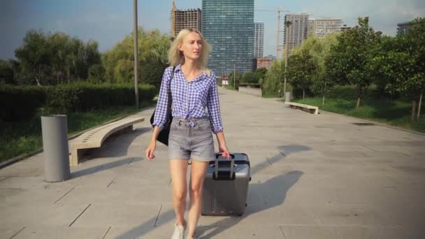 Молодая красивая женщина проходит через курортный город и катит чемодан позади нее. — стоковое видео