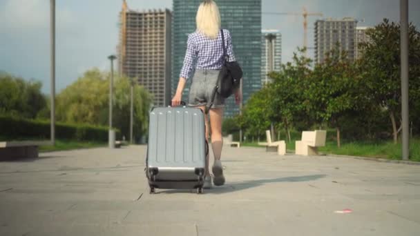 Joven hermosa mujer camina a través de ciudad turística y rueda maleta detrás de ella. — Vídeos de Stock