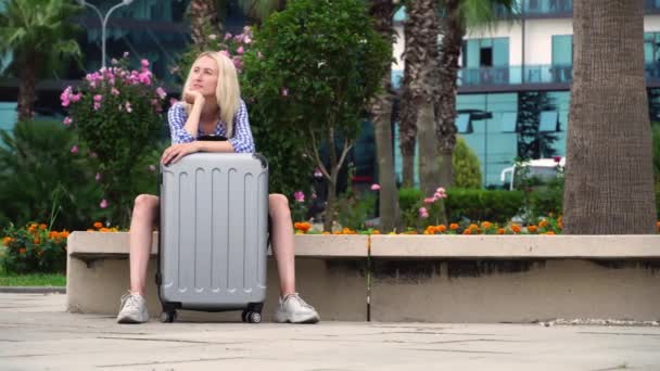 Eine hübsche Frau mit grauem Koffer sitzt auf einer Parkbank. ein Smartphone. — Stockvideo