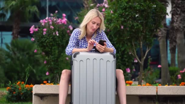 Una mujer guapa con una maleta gris está sentada en un banco en el parque. un smartphone. — Vídeos de Stock