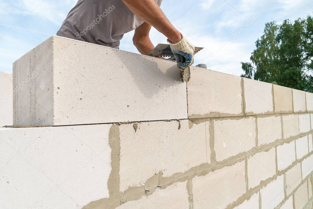 Worker wipes the seams with a spatula outside the house of blocks. Bricklayer worker laying white blocks of a future house in the open air.