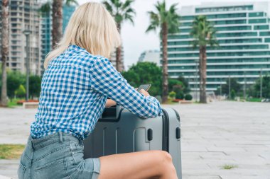 İnce, mavi tişörtlü, gri kot şortlu genç, sarışın bir kadın cep telefonuyla bankta oturuyor ellerini gri bir bavula yaslıyor ve bacaklarının arasında duruyor.