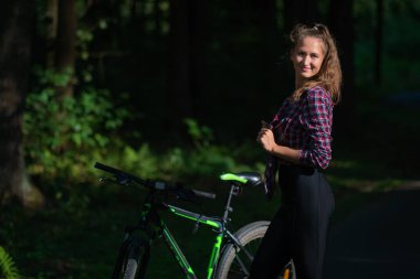 Güneşli bir yaz gününde, tişörtlü, taytlı ve bisikletli gülümseyen genç bir kadının portresi.