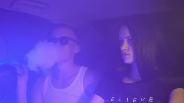 Una giovane coppia piacevole sta fumando sul sedile posteriore di un'auto in una luce al neon. — Video Stock