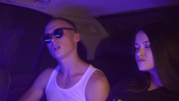 Egy fiatal, kellemes pár dohányzik egy autó hátsó ülésén egy neonfényben.. — Stock videók
