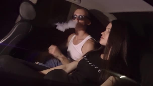Una joven pareja agradable está fumando en el asiento trasero de un coche en una luz de neón. — Vídeos de Stock