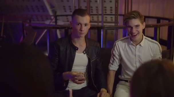 Rapazes e raparigas relaxam numa mesa numa discoteca. Eles riem, bebem, falam . — Vídeo de Stock