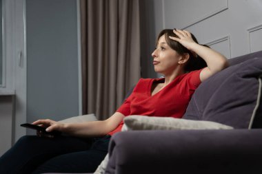 Düşünceli bir kadın, elinde uzaktan kumandayla rahat bir koltukta oturuyor ve televizyon içeriğini yansıtıyor, evinde bir anlık tefekkür ve rahatlamaya dalmış.