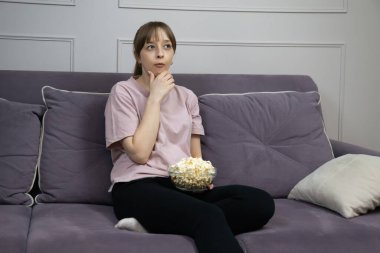 Düşünceli genç bir kadın TV 'de film izliyor, rahat bir koltukta oturuyor ve elinde patlamış mısırla evde boş zamanlarının keyfini çıkarıyor.