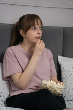 Genç bir kadın yatakta rahat bir şekilde oturur, bir filme dalmış ve bir kase patlamış mısırın tadını çıkararak evinde rahatlatıcı bir akşamın ruhunu yakalar. Dikey fotoğraf