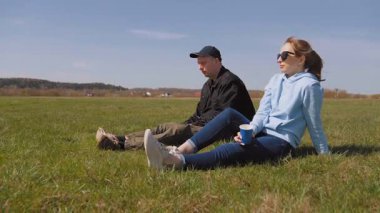 Genç bir adam ve kadın güneşli yeşil bir alanda oturuyor, dinleniyor, konuşuyor ve güzel bahar gününün tadını çıkarıyor, barışçıl bir piknik sırasında sohbet ediyor ve doğanın tadını çıkarıyorlar.
