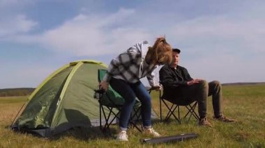 Genç bir adam ve kadın güneşli bir günde güzel manzaranın ve temiz havanın tadını çıkarmak için çadırlarının yanındaki güneşli çayırda kamp sandalyelerini birleştiriyorlar.