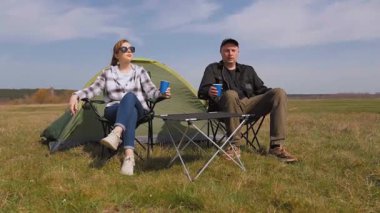 Kafkasyalı rahat bir çift çadırın yanındaki kamp sandalyelerinde oturur ve kupalardan içerler. Güneşli güzel günün ve kırsal alandaki maceralarının tadını çıkarırlar.