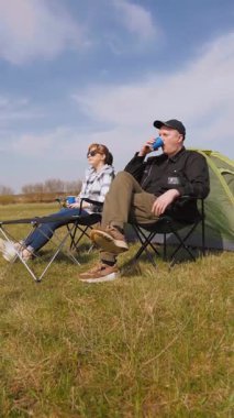 Kafkasyalı rahat bir çift çadırın yanındaki kamp sandalyelerinde oturur ve kupalardan içerler. Güneşli güzel günün ve kırsal alandaki maceralarının tadını çıkarırlar.