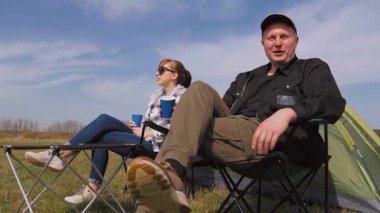 Kafkasyalı rahat bir çift çadırın yanındaki kamp sandalyelerinde oturur ve kupalardan içerler. Güneşli güzel günün ve kırsal alandaki maceralarının tadını çıkarırlar.