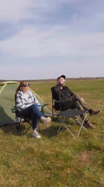 Kafkasyalı rahat bir çift çadırın yanındaki kamp sandalyelerinde oturur ve kupalardan içerler. Güneşli güzel günün ve kırsal alandaki maceralarının tadını çıkarırlar.