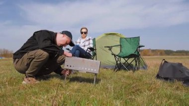 Genç bir adam çimenli bir alanda piknik yapmak için portatif barbekü ızgarası yaparken kız arkadaşı da yakınlardaki katlanır sandalyede dinleniyor ve birlikte açık hava maceralarının tadını çıkarıyor.