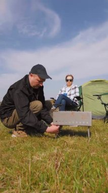 Genç adam çimenli bir alanda piknik yapmak için portatif barbekü ızgarası yapıyor. Kız arkadaşı da yakınlardaki katlanır sandalyede dinleniyor ve açık hava maceralarının keyfini çıkarıyor.